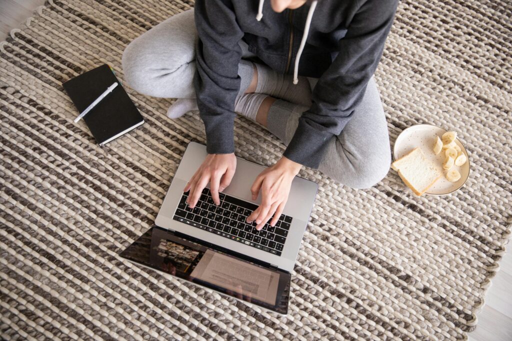 pexels-photo-4049730-4049730 Woman in casual wear working remotely on a laptop, with a notebook and snack nearby.
